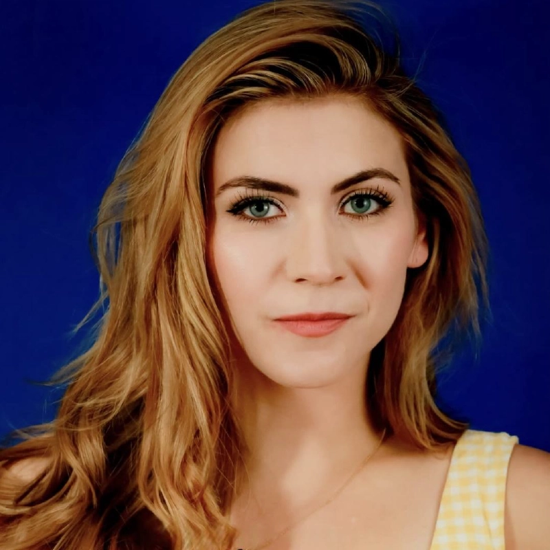 A woman with long, wavy light brown hair and blue-green eyes stares directly at the camera. She wears a yellow plaid sleeveless top and a gold necklace. The background is a solid deep blue, and her expression is neutral and confident.