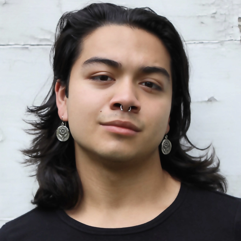 A person with long dark hair, wearing silver earrings and a septum piercing, poses in front of a white textured wall. They have a neutral expression and are wearing a black shirt.