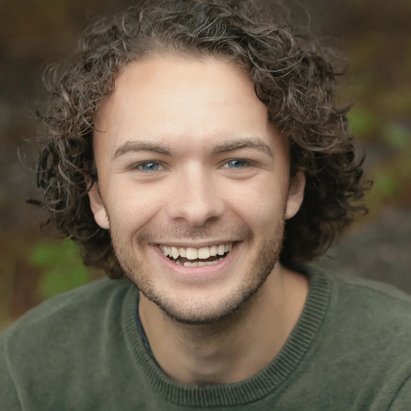 A young man with fair skin, curly brown hair, blue eyes, and a short beard smiles broadly at the camera. He is wearing a green sweater and is outdoors, with a softly blurred natural background.