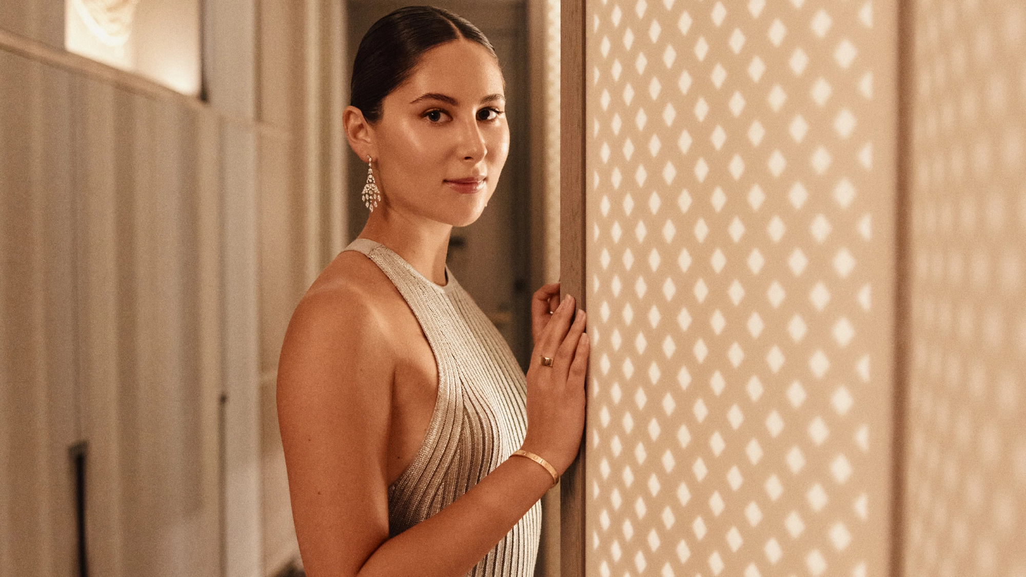 A woman with dark hair in a sleek bun stands indoors, wearing a shimmery silver dress and dangling earrings. She looks at the camera, gently touching a beige wall with a diamond light pattern, as if posing for an album cover by Eden Plepler.
