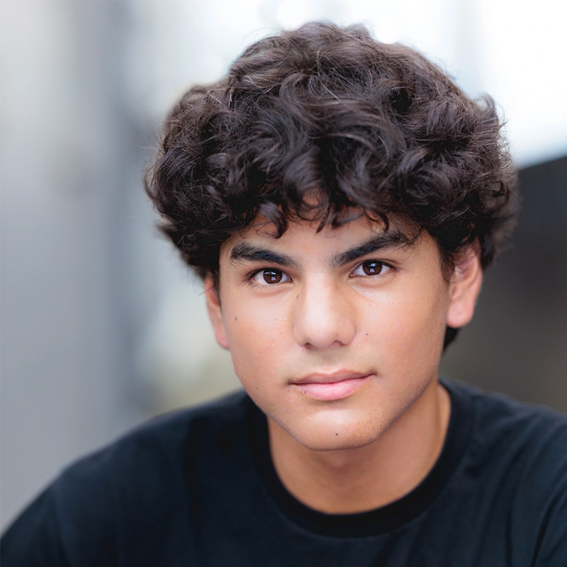 A young person with curly dark hair and light brown skin looks directly at the camera with a neutral expression. They are wearing a plain black shirt. The softly blurred background in tones of gray and white adds focus to Sophia Leverett's gaze.
