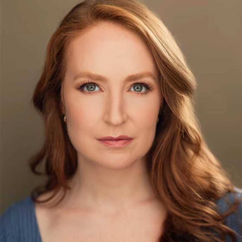 Ellie Fishman, a woman with long, wavy red hair and blue eyes, gazes directly at the camera. She has a fair complexion, natural makeup, and wears a blue top. The softly blurred warm background highlights her striking features.