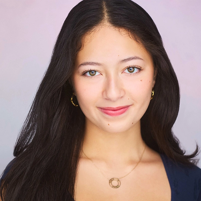 Addison Wivagg, a young woman with long dark hair, green eyes, and light skin, smiles gently at the camera. She wears a navy top, gold hoop earrings, and a circular pendant necklace against a soft, light background that highlights her face.