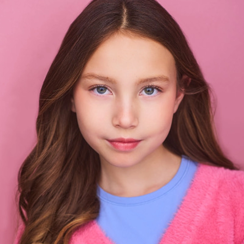 A young girl with long, wavy brown hair and blue eyes looks at the camera. She wears a light blue shirt under a pink fuzzy sweater. The solid pink background creates a soft, colorful portrait by Jackie Winslow; her expression is calm and neutral.