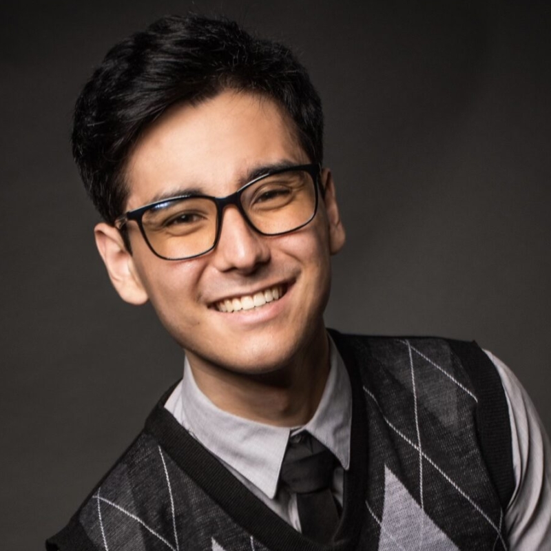 A young man with short dark hair and glasses smiles at the camera. He is wearing a gray argyle sweater vest over a light shirt and dark tie. The background is dark and plain, highlighting his friendly expression and professional attire.
