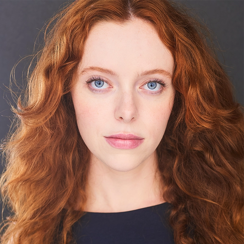 A woman with long, wavy red hair and pale skin looks directly at the camera. She has blue eyes, light makeup, and a neutral expression. Reminiscent of a Timothy Thomas portrait, she wears a dark top against a plain, dark background.