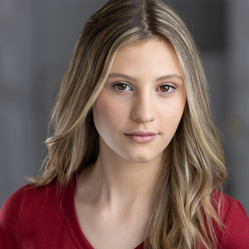 A young woman with long, light brown hair and fair skin looks directly at the camera. Sophia Leverett wears a red top and subtle makeup, with a softly blurred background in neutral tones highlighting her gentle expression.