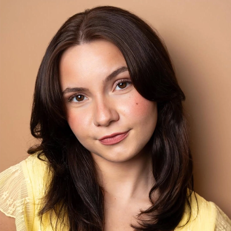 Emily Rose DeMartino, a young woman with medium-length dark brown hair and fair skin, smiles softly. She wears a yellow lace-trimmed top and subtle makeup on her cheeks and lips. The background is a solid, warm light brown color.