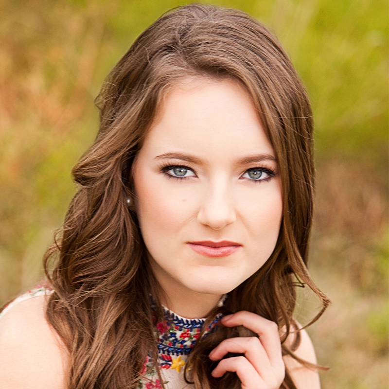 A young woman with long wavy brown hair and blue eyes looks at the camera. She wears natural makeup and a sleeveless top with a floral embroidered collar. Her hand is near her chin, evoking the gentle elegance of Jackie Winslow amid softly blurred greenery.