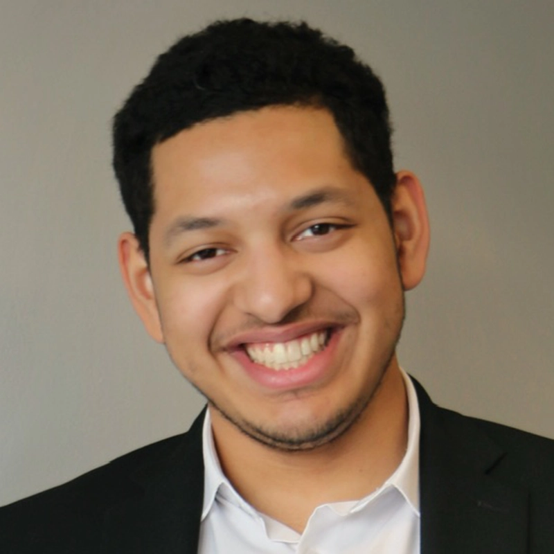 Emmanuel Sacasa Reyes, a young man with short, curly black hair and trimmed facial hair, smiles broadly. He is wearing a black blazer over a white collared shirt against a plain gray background, giving the image a professional, portrait-like appearance.