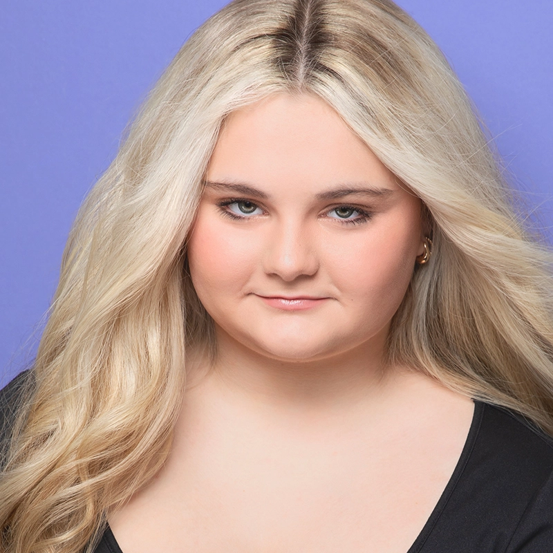 A young woman, Eva Joan Daugherty, with long, wavy platinum blonde hair and fair skin looks directly at the camera. She is wearing a black top and small hoop earrings. The background is solid light purple; she has a subtle smile and natural makeup.