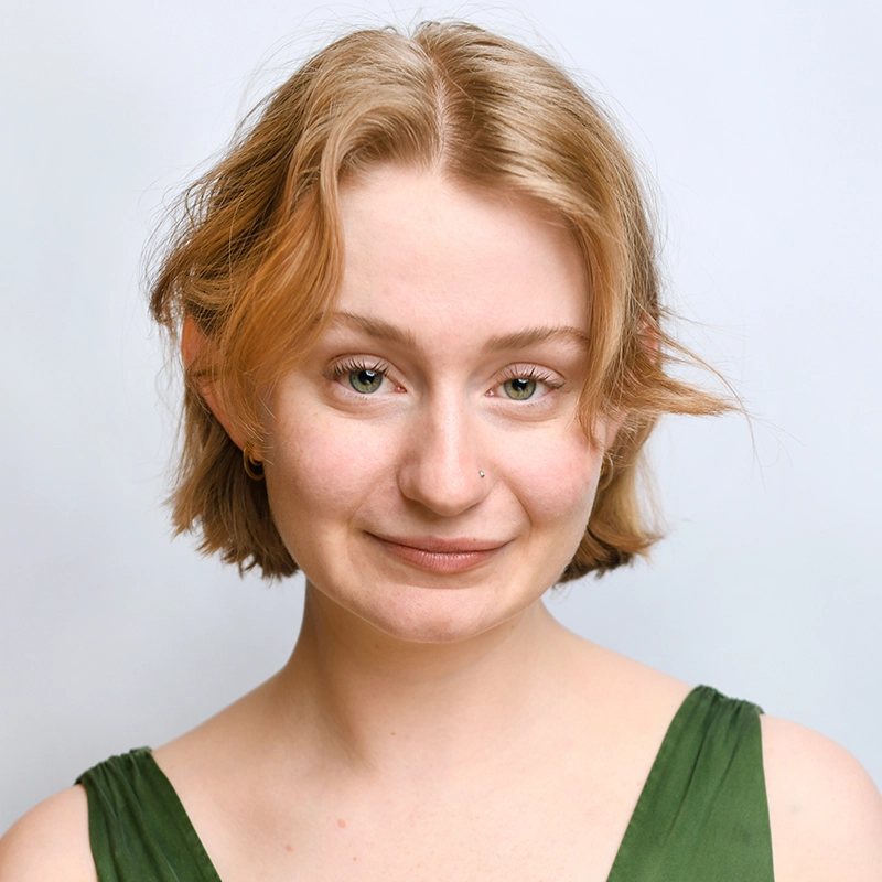 A young person with short, light brown hair and fair skin smiles softly at the camera. Maggie Weller has a small nose piercing and wears a green sleeveless top, set against a plain, light-colored background that keeps the focus on her face.
