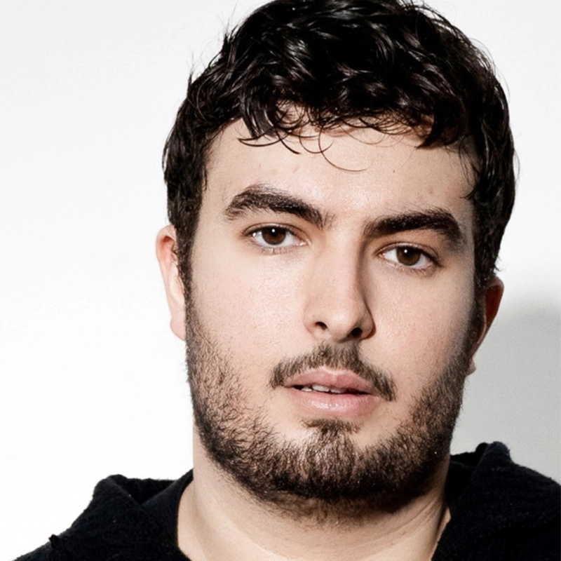 A young man with short, dark, wavy hair and a trimmed beard looks at the camera with a neutral expression. He is wearing a black hoodie, photographed by Maggie Weller, and stands against a plain white background.