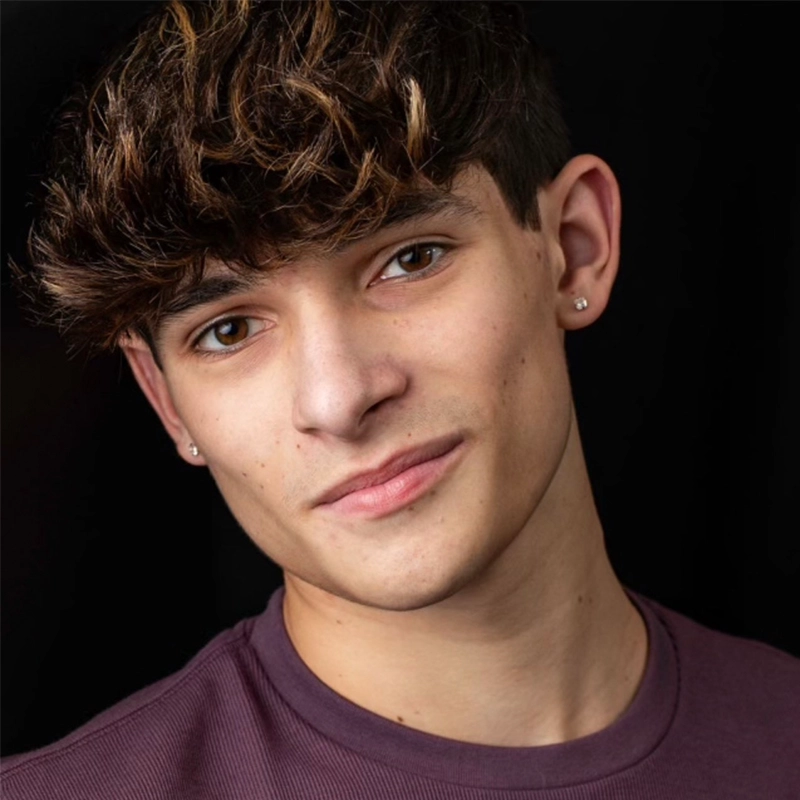 Shayla Lopez, a young person with short, wavy brown hair and fair skin, wears a maroon shirt. Small stud earrings accent both ears as Shayla offers a slight smile. The black background and lighting highlight her facial features.