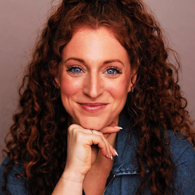 Dara Gallagher, with long, curly red hair, smiles warmly at the camera. Her bright blue eyes and light skin glow as she poses thoughtfully in a denim jacket against a soft, neutral background.