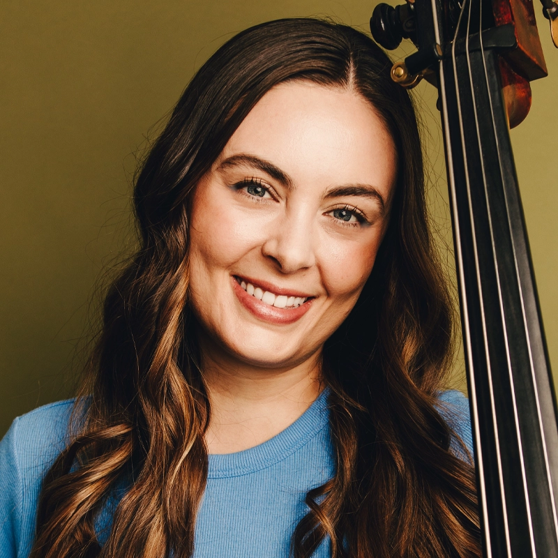 Lauren Gamiel, a woman with long, wavy brown hair, smiles warmly at the camera. She wears a light blue top and stands next to the neck of an upright bass against a soft olive green background.