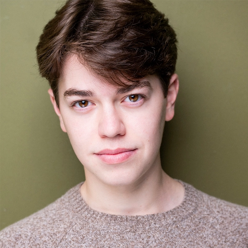 Sophia Leverett, a young person with short brown hair and fair skin, looks directly at the camera in a beige knit sweater. The plain olive green background and soft lighting highlight her gentle expression and facial features.