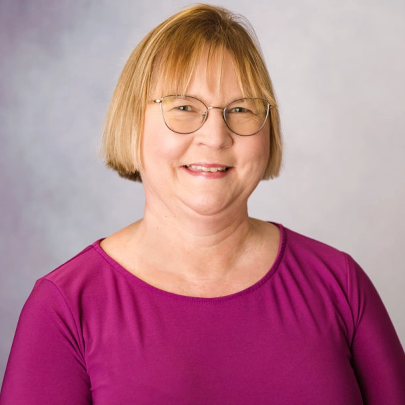 Ann Gerschefski, a woman with short blond hair and glasses, smiles at the camera. She is wearing a magenta top and stands against a soft, light gray background.