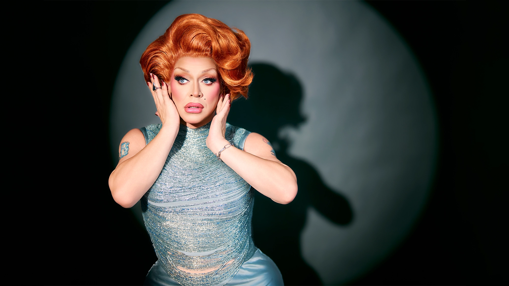A drag queen with voluminous orange hair, dramatic makeup, and a shimmering silver-blue sleeveless gown poses with hands framing their face, looking surprised. A spotlight casts their shadow on a dark background.