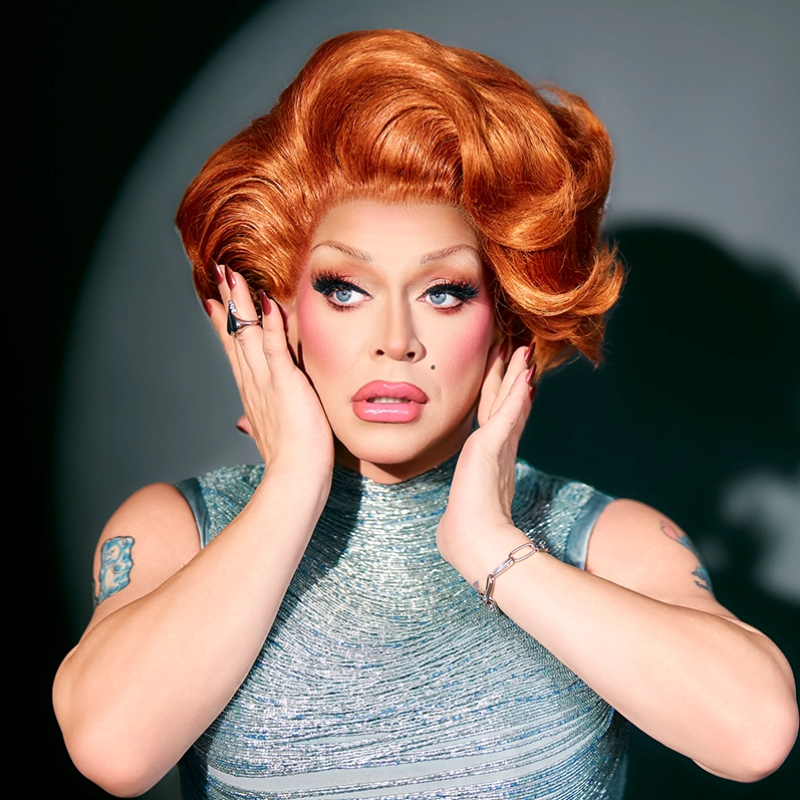A person in glamorous drag with voluminous red hair, bold makeup, and dramatic false eyelashes, wears a shiny silver sleeveless top. Their hands are raised near their face in a surprised pose against a dark, blurred background.