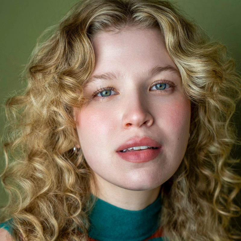A young woman with pale skin and wavy blonde hair looks at the camera with a neutral expression. She has blue eyes, pink lips, and wears a green high-neck top. The softly blurred green background highlights Isabel Ginsberg’s calm presence.