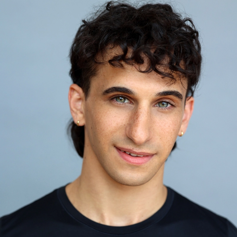 Dani Goldberg, with short, curly dark hair and green eyes, smiles softly at the camera. They wear small gold hoop earrings and a black shirt against a plain, light gray background.
