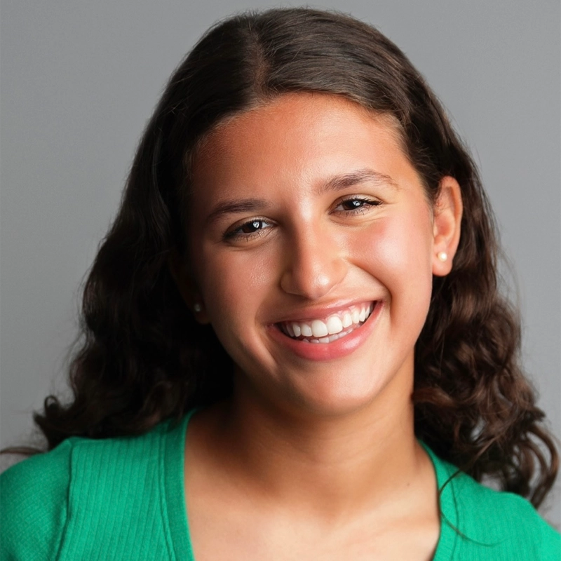 Tali Goldwasser, a young woman with long, wavy brown hair, smiles warmly at the camera. She is wearing a green top and small stud earrings, with a plain gray background emphasizing her friendly expression and natural look.