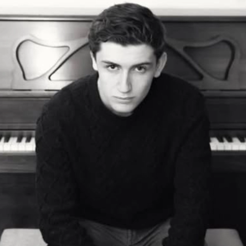 A young man with dark hair, wearing a textured dark sweater, sits in front of an upright piano. Joshua Gorlin looks directly at the camera with a serious expression in this black and white photo, emphasizing contrasts in his face and surroundings.