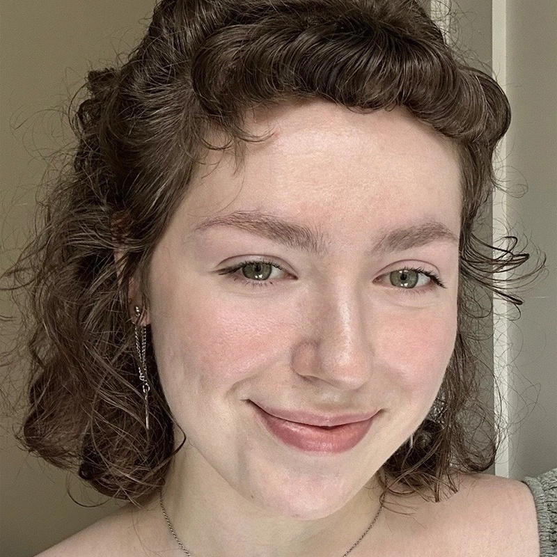 A young person with fair skin, short curly brown hair, and green eyes smiles gently at the camera. Styled like Giovanna Greco, they wear subtle pink makeup, delicate earrings, and a thin necklace against a softly lit, neutral background.