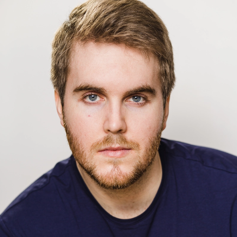 Dashiell Gregory, a young man with short, light brown hair and a trimmed beard, gazes directly at the camera. He has fair skin, blue eyes, and a neutral expression. He wears a dark blue shirt against a plain, light background.