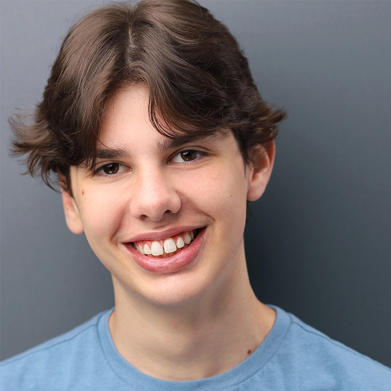 A teenage boy with light skin and brown, wavy hair smiles at the camera. Wearing a light blue T-shirt, he stands against a soft gradient gray background, creating a friendly and approachable portrait captured by Shayla Lopez.