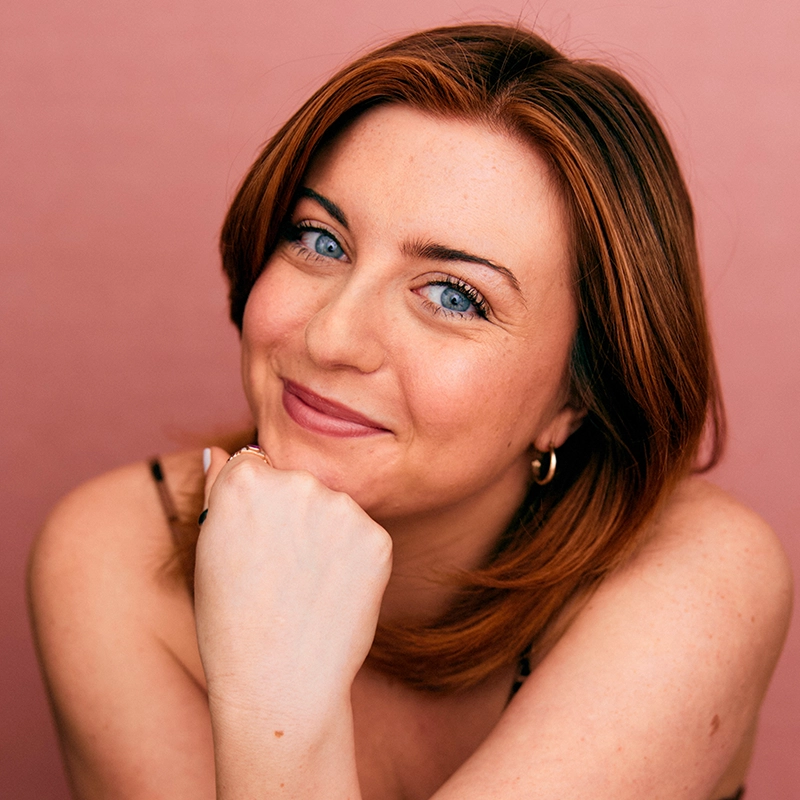 A person with fair skin, shoulder-length auburn hair, and blue eyes smiles softly. They rest their chin on their hand and wear a sleeveless top and hoop earrings. The background is a solid pink color, creating a warm and friendly atmosphere.