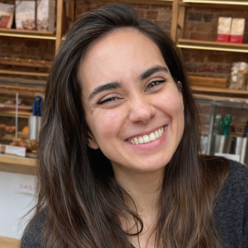 Emily Harrington, a young woman with long brown hair, smiles warmly at the camera. She is indoors, possibly in a café or bakery with shelves of baked goods and brick walls in the background, creating a cozy and inviting atmosphere.