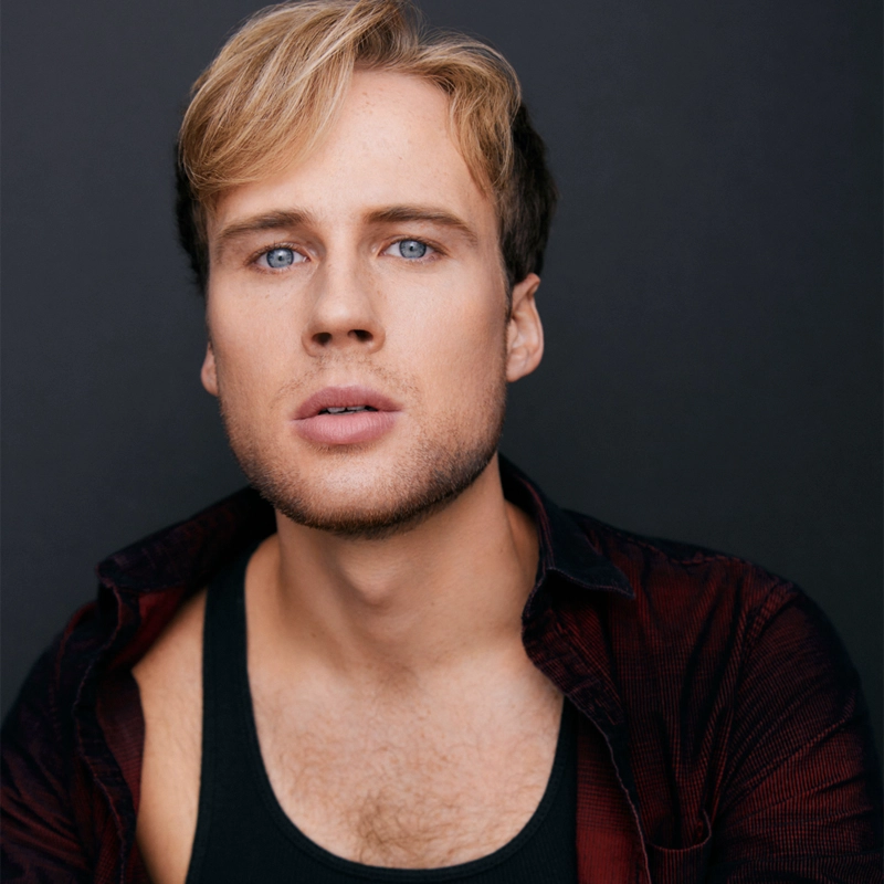 Charlie High, a young man with light skin, short blonde hair, and blue eyes, looks directly at the camera. He has light facial hair and is wearing a black tank top under a dark red open shirt against a plain, dark background.