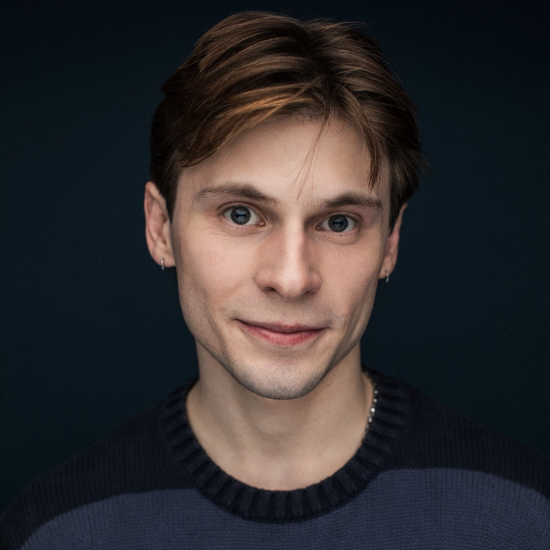 A young man with fair skin and light brown hair looks at the camera, smiling softly. He is wearing a dark sweater and small hoop earrings in both ears. The background is dark, drawing focus to his face.