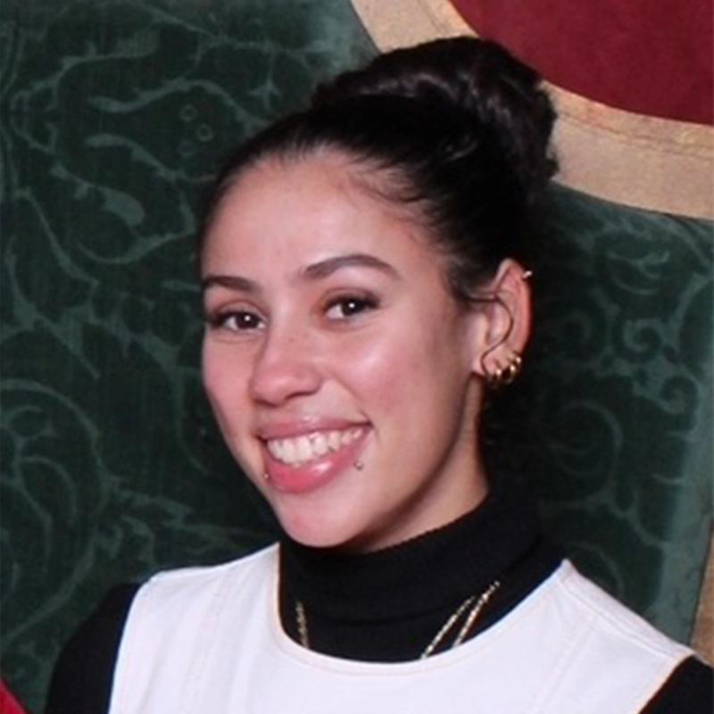 Isabella Cruz, a young woman with dark hair tied in a bun, smiles, showing a lip piercing. She wears hoop earrings, layered necklaces, a black turtleneck, and a white sleeveless top against green patterned fabric with touches of red and gold.