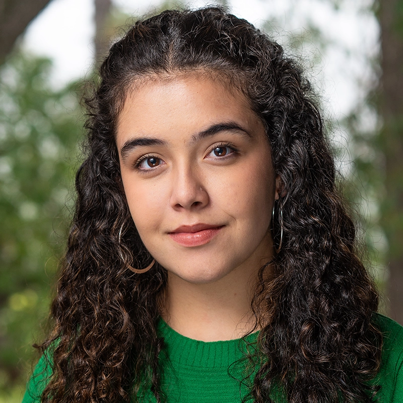 A young woman with long, curly dark hair and hoop earrings looks at the camera with a slight smile. She is wearing a green sweater, outdoors near softly blurred trees—an NYU student with a passion for mariachi music shining through.