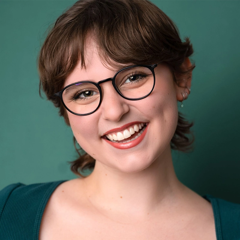 Sophia Leverett, a young woman with short brown hair and glasses, smiles warmly at the camera. She is wearing a teal top and small earrings, standing against a solid green background. Her expression is cheerful and inviting.