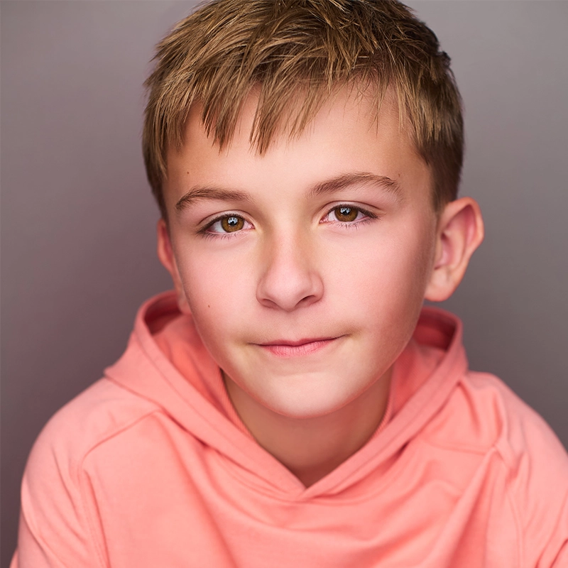 A young boy with short, light brown hair and hazel eyes wears a peach-colored hoodie. Jackie Winslow looks directly at the camera with a slight, closed-mouth smile. The neutral gray background makes his face and clothing stand out clearly.