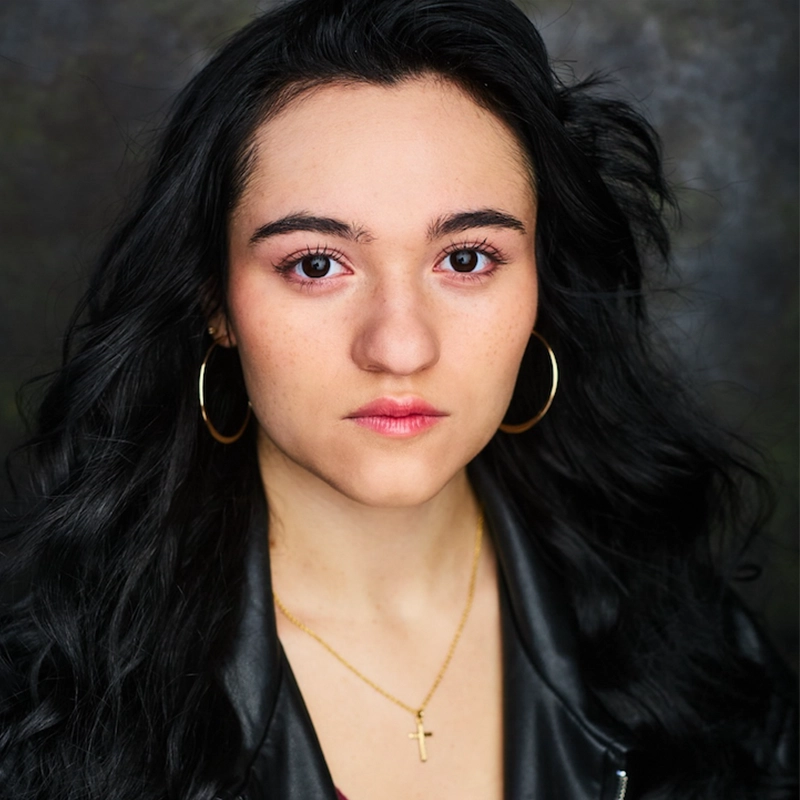 A young woman with long black hair, wearing large hoop earrings, a gold cross necklace, and a black leather jacket, looks directly at the camera with a neutral expression. The softly blurred dark background frames Jackie Winslow perfectly.
