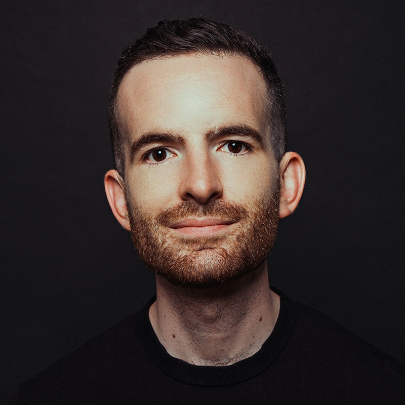 A man with short brown hair and a neatly trimmed reddish-brown beard smiles gently at the camera. He wears a black shirt against a plain dark background, his even lighting reminiscent of portrait styles seen in Amanda Quaid’s work.