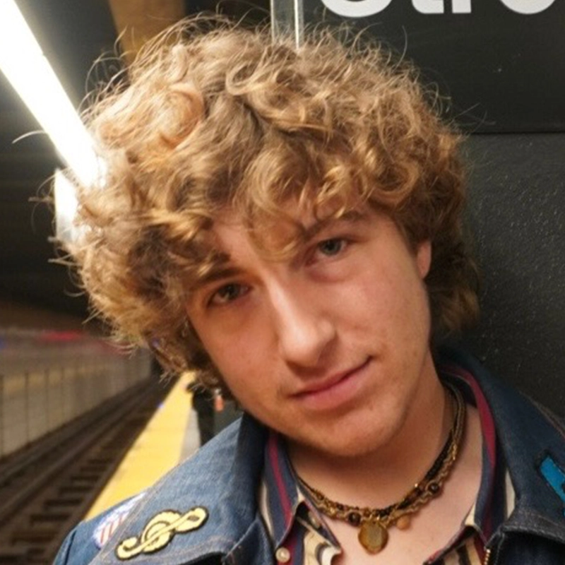 A young person with curly blonde hair and layered necklaces looks into the camera, standing on a subway platform beside the tracks. They wear a denim jacket with a musical note patch. The station’s tile wall and overhead lights are visible in the background.