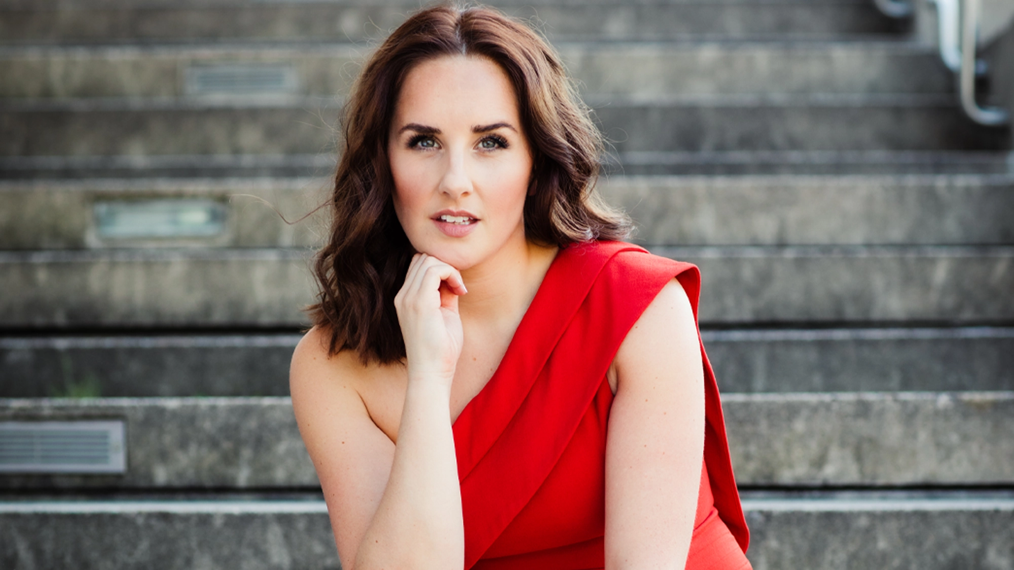 A woman with wavy brown hair wearing a one-shoulder red dress sits on stone steps, gazing at the camera with a slight smile. Inspired by Songs I Love to Sing, she exudes the poised charm of Jeanna de Waal. The background is softly blurred.