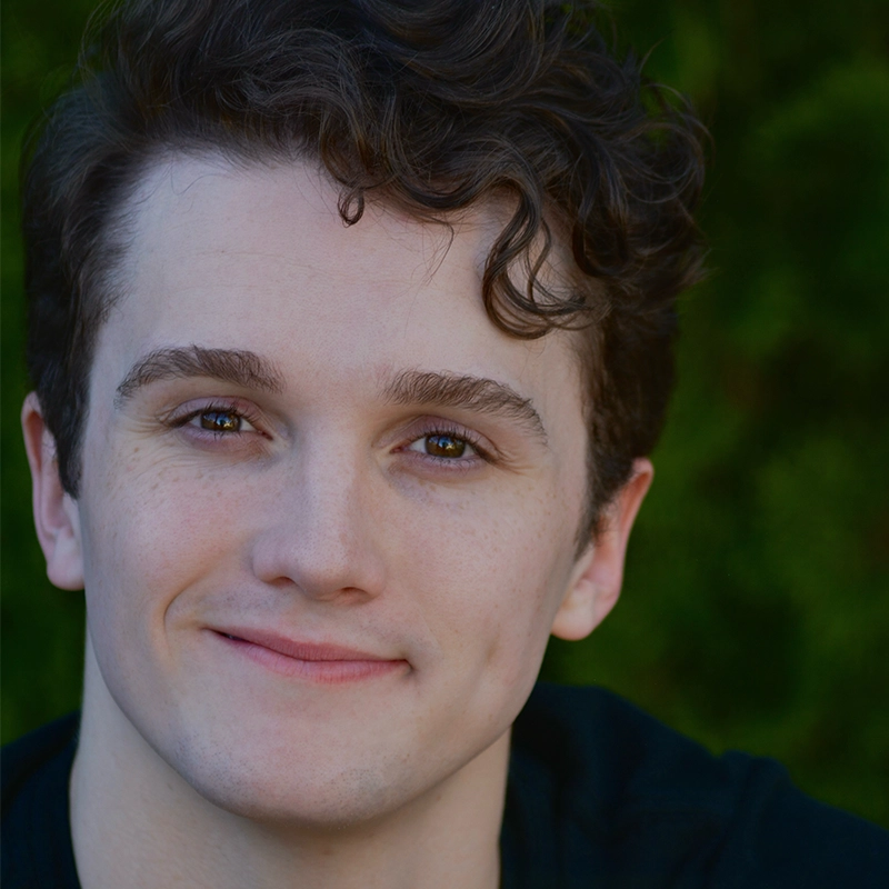 A young person with light skin and short, curly brown hair smiles gently at the camera. Their eyebrows are dark and full, and they have brown eyes. The background is blurred green foliage, suggesting an outdoor setting.