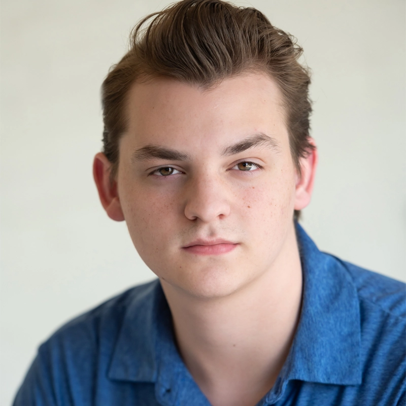 Zachary Johnson, a young man with light skin and short, swept-back brown hair, looks directly at the camera in a blue collared shirt. He has a neutral expression and is set against a plain, light background.