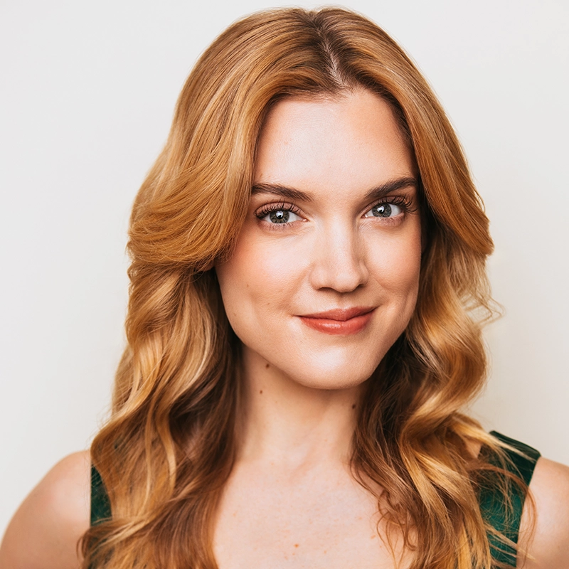 Alice K. Tinari, with wavy strawberry blonde hair and fair skin, smiles gently. She has light makeup, brown eyes, and wears a sleeveless green top. The plain white background keeps the focus on her face.
