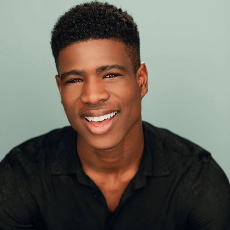 A young man with short, curly black hair smiles at the camera. He wears a black button-up shirt and sits against a light teal background. His expression is friendly and approachable, with his teeth showing as he smiles.