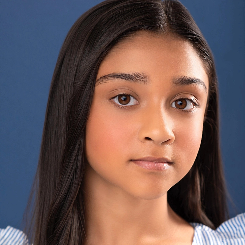 A young girl, Luli Mitchell, with long, straight dark hair and brown eyes looks at the camera with a neutral expression. She has smooth, light brown skin and wears a blue and white top. The background is solid dark blue.