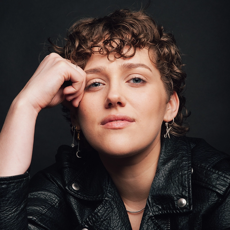 Maggie Weller, with short, curly brown hair and blue eyes, wears a black leather jacket and silver earrings, posing confidently with her head resting on her hand against a dark background. She has a relaxed expression and a slight smile.