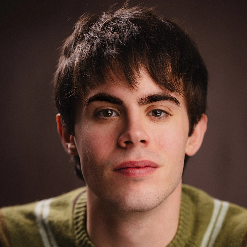 A young man with short brown hair and fair skin looks directly at the camera, wearing a green sweater with white stripes on the shoulders. The background is softly blurred, focusing attention on his calm, neutral expression.